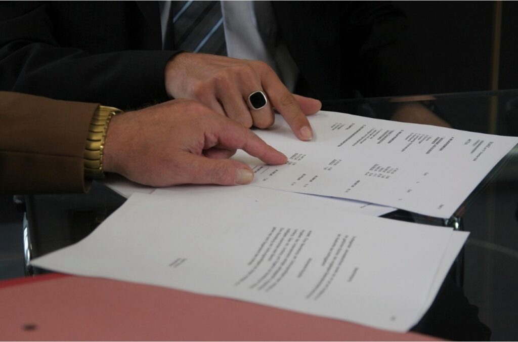 Close-up of two people's hands reviewing printed documents on a glass table, with one hand wearing a black signet ring pointing at the paper and the other wearing a gold bracelet.