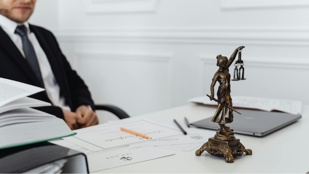 Bronze Lady Justice statue on desk with legal documents, laptop, and a professional in formal attire.