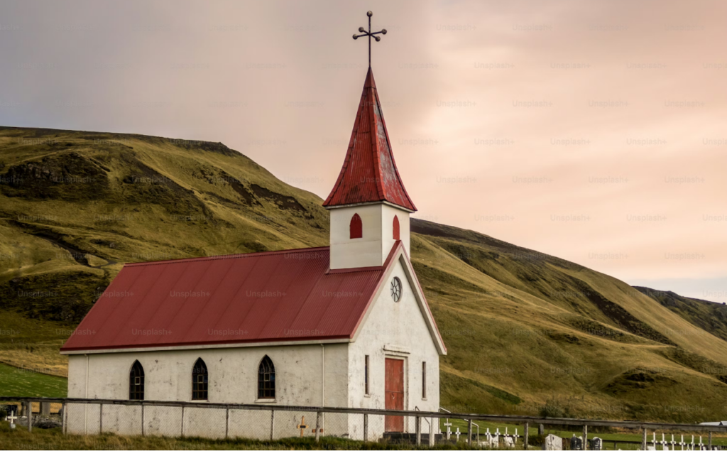 White church with red roof and steeple against green hillside with cemetery in foreground