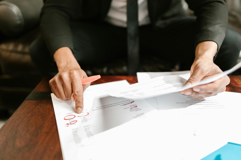 Person in dark clothing reviewing documents with red markings and highlights on a wooden table.