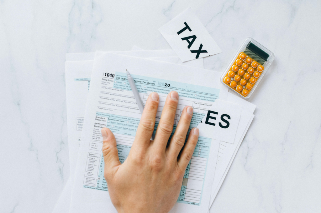 Hand resting on Form 1040 tax documents with "TAX" label and orange calculator on marble surface.