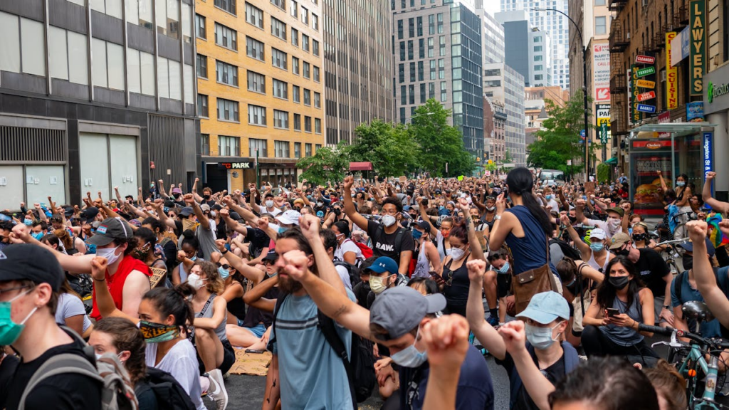 Protesters marching through a city, surrounded by tall buildings and trees.