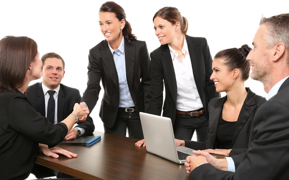 Group of six smiling business professionals in dark suits gathered around a wooden conference table with a laptop and smartphone, with two women shaking hands across the table.