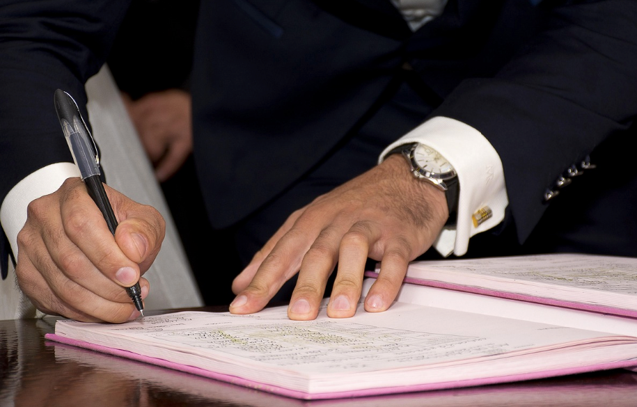 Close-up of a man in a dark suit, white dress shirt, and silver wristwatch signing a document in a pink folder with a black pen at a desk.Â