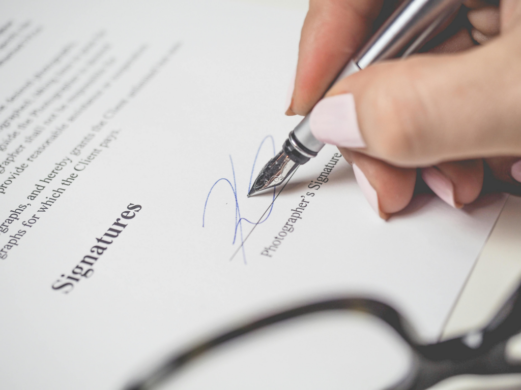 A hand signing a document with a fountain pen, with glasses resting nearby.