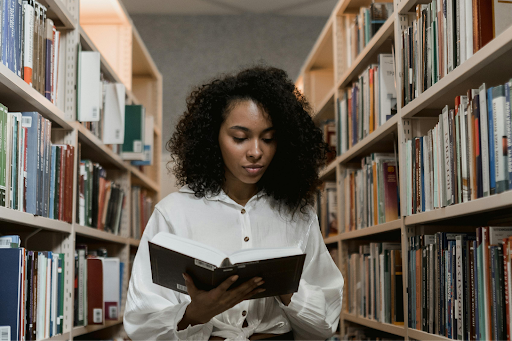 A woman with curly hair reads a book while standing between library shelves.