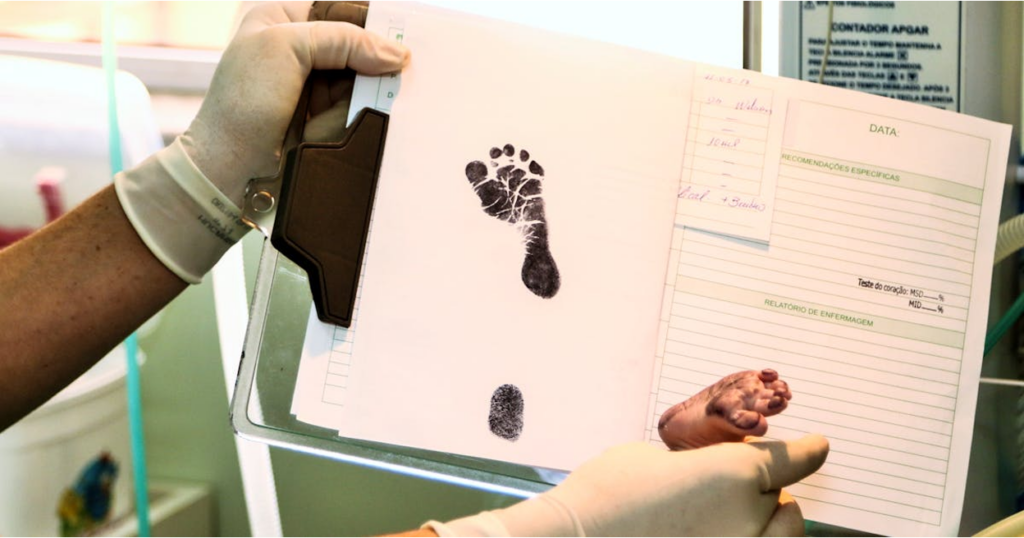 Gloved hands holding a newborn's medical record with an ink footprint and a tiny baby foot visible