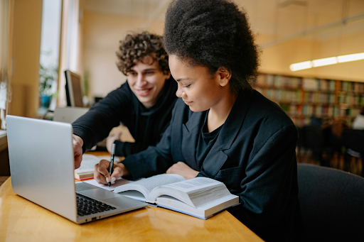 Two students study together at a library table with a laptop and an open textbook.