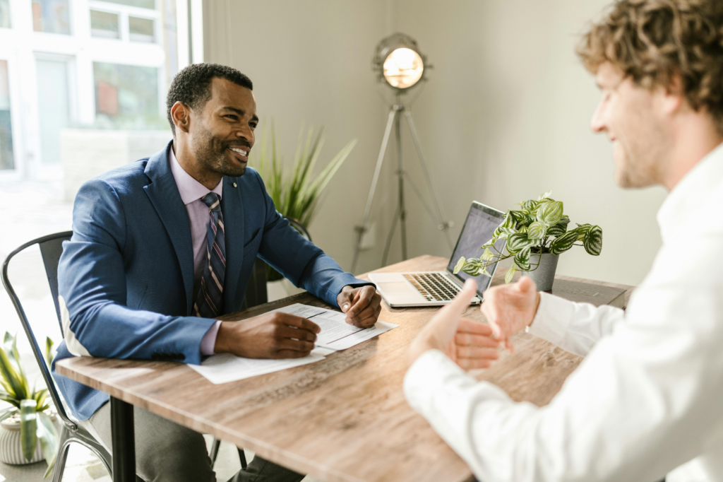 Two men in business attire having a friendly conversation at a desk with a laptop.