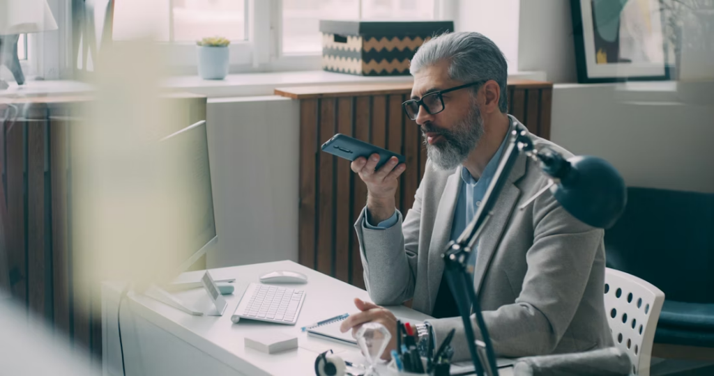 A man wearing glasses talks on the phone while working at a desk with a computer.