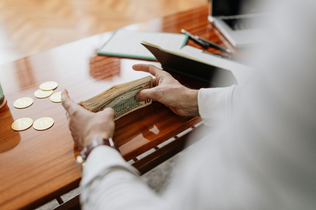 Hands counting a stack of US dollar bills on a wooden desk with gold coins nearby.