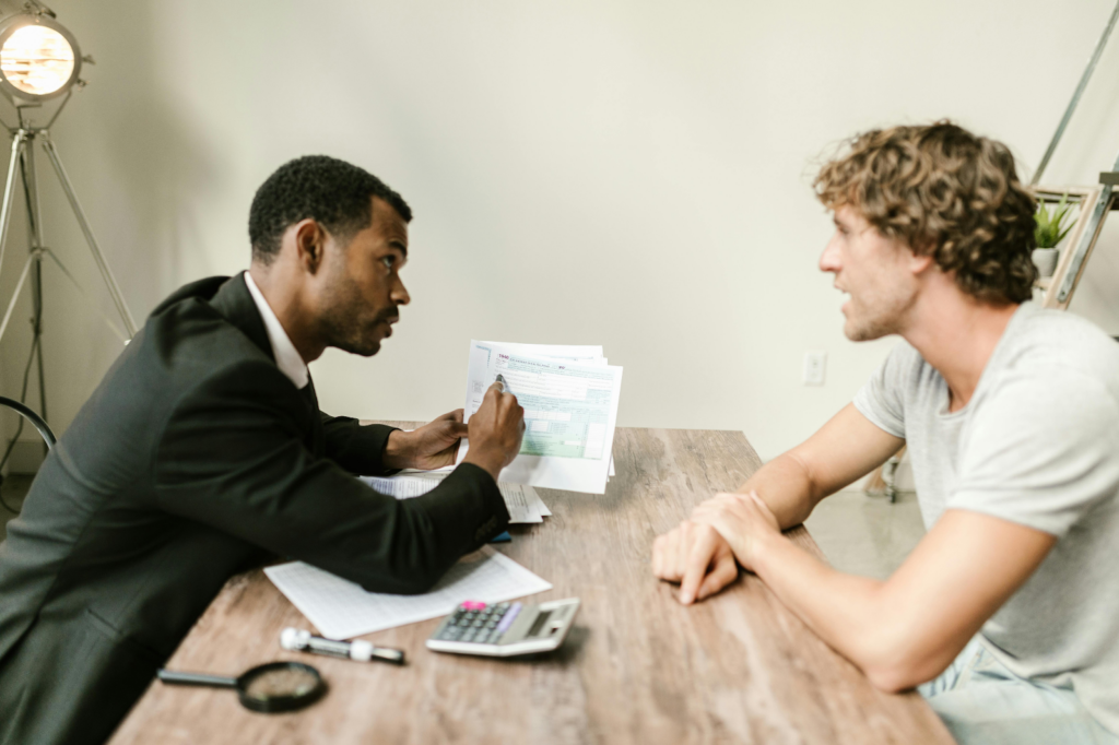 A man in a suit shows tax documents to another man across a wooden desk with a calculator.