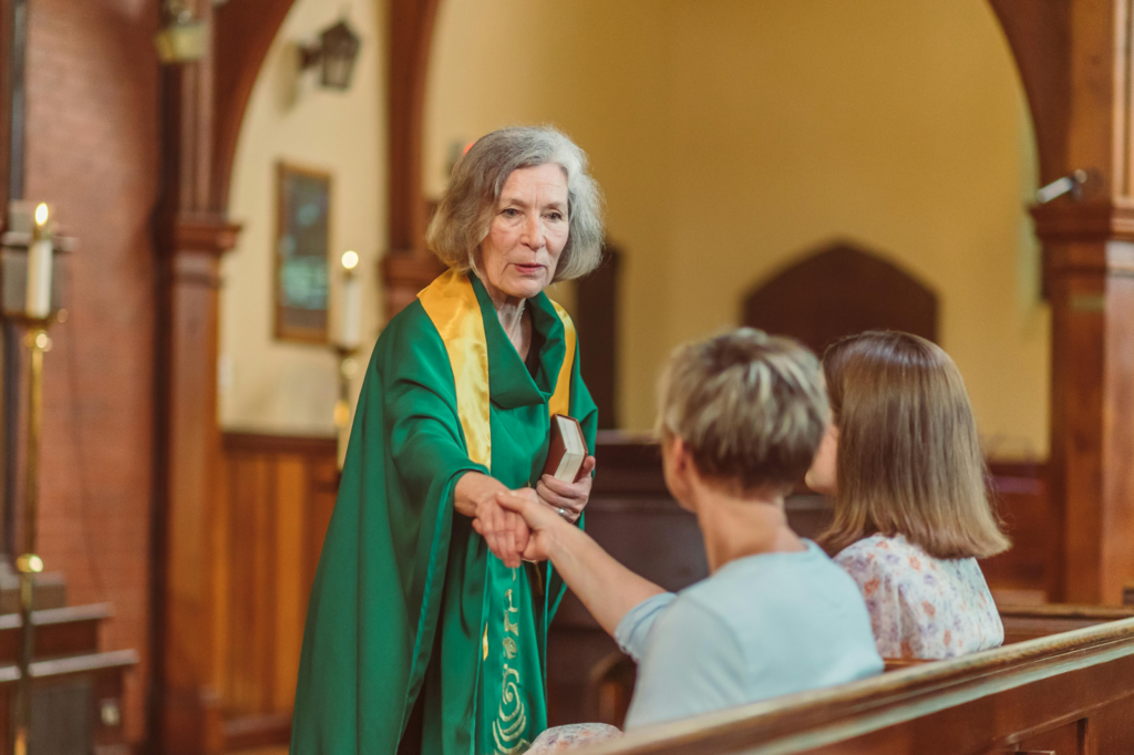 A clergy member in green vestments shaking hands with parishioners seated in church pews.