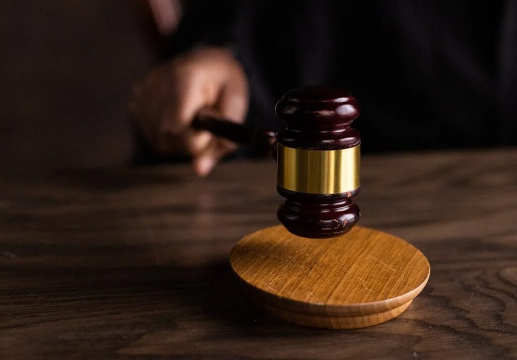 A judge's hand striking a wooden gavel on a sound block on a dark wooden surface.
