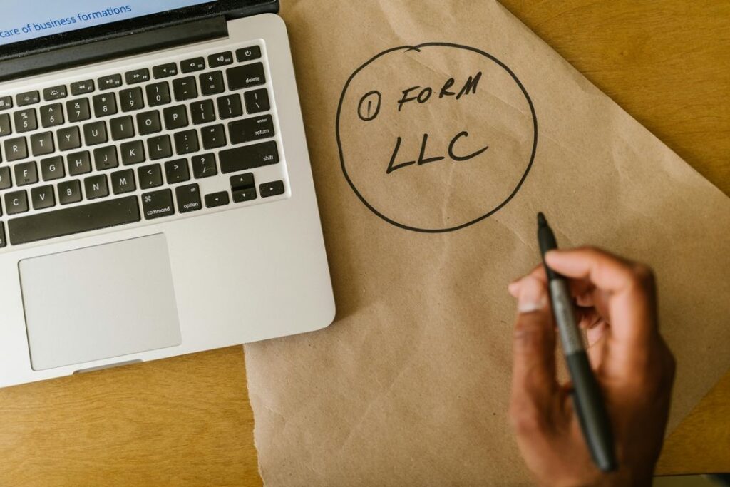 A hand writing "Form LLC" on brown paper next to an open laptop on a desk.