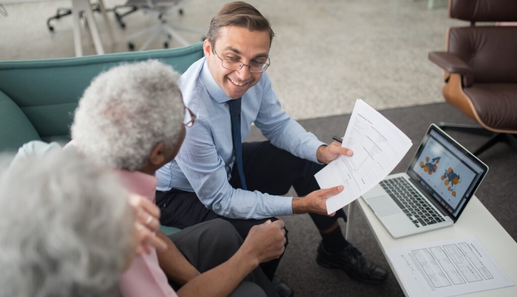 A smiling financial advisor showing documents to an elderly couple beside an open laptop.