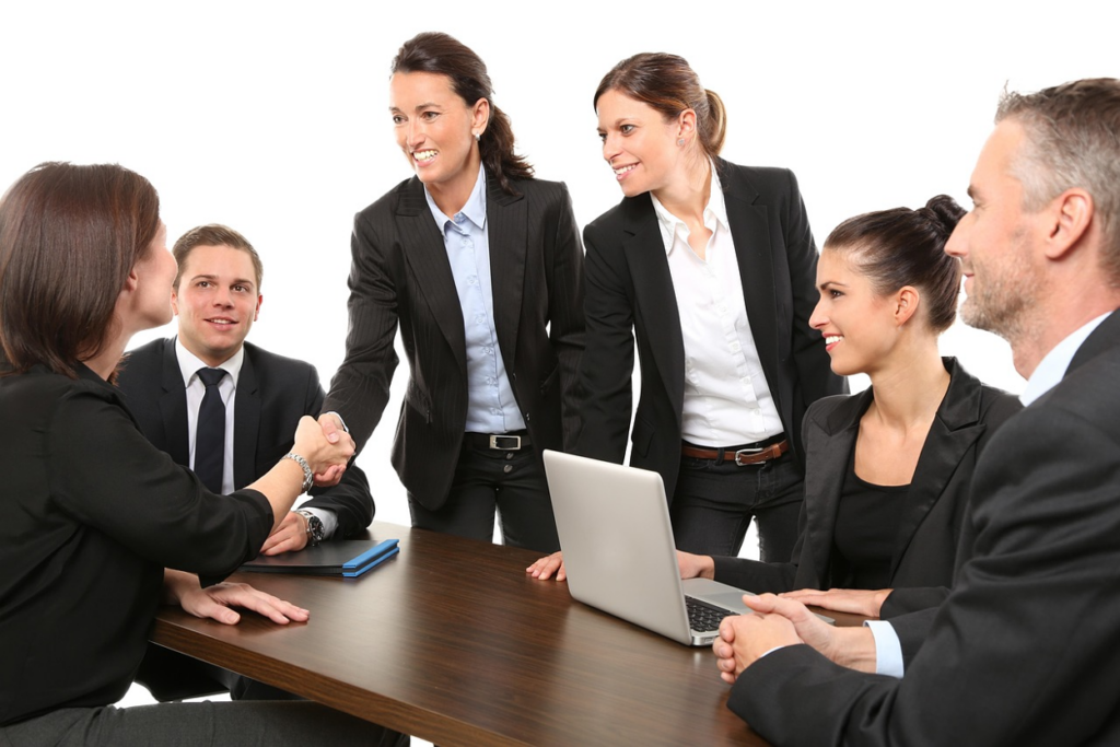 Six business professionals in formal attire engaged in conversation in an office with two of the associates having a handshake