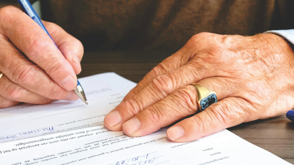 An elderly person's hands signing a legal document with a fountain pen on a desk.