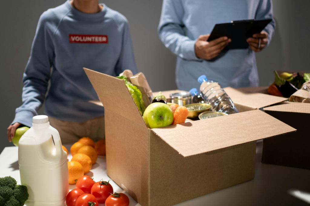Two volunteers in blue sweatshirts are packing a donation box with fresh produce, canned goods, and water.