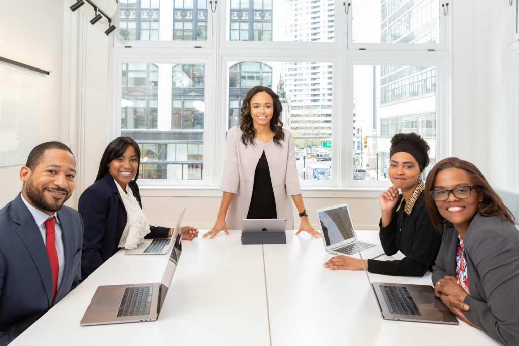 Five professionals in business attire smiling around a conference table with laptops in a bright city office.
