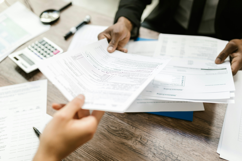 Two people exchanging tax documents across a wooden desk with a calculator, pen, and paperwork.