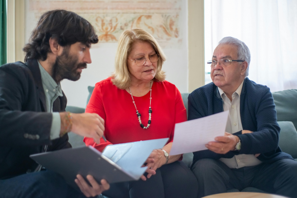 A young man shows documents to an older couple seated on a couch during a consultation.