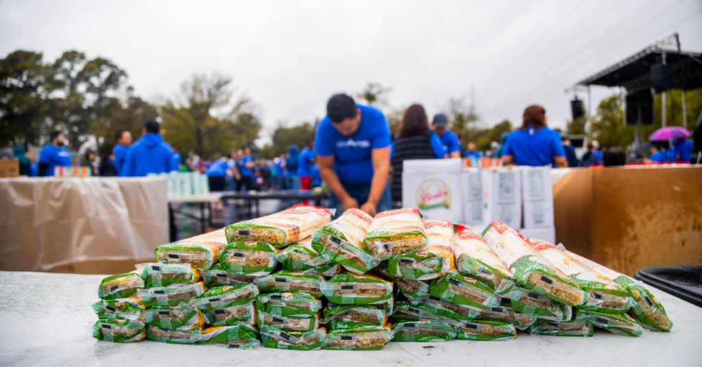 Volunteers in blue shirts organize stacked bags of pasta at an outdoor charity food distribution event.