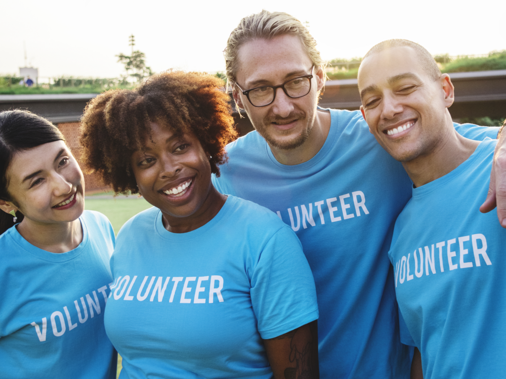 Four smiling volunteers in blue "Volunteer" t-shirts pose together outdoors.