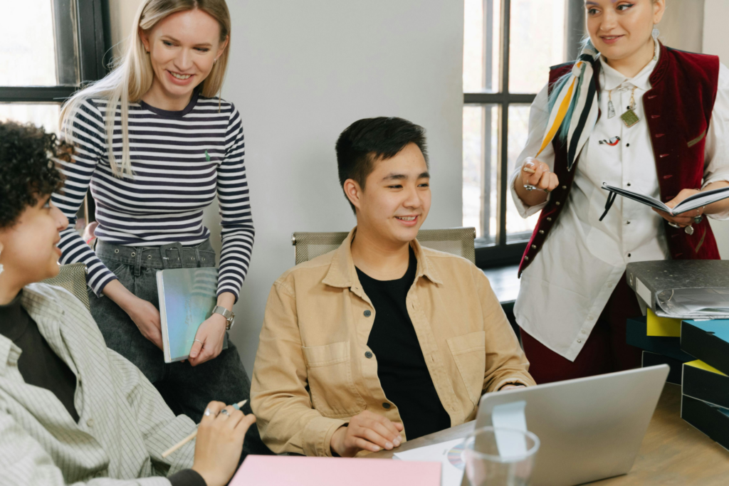 Four young colleagues smile and collaborate around a laptop in a bright office setting.