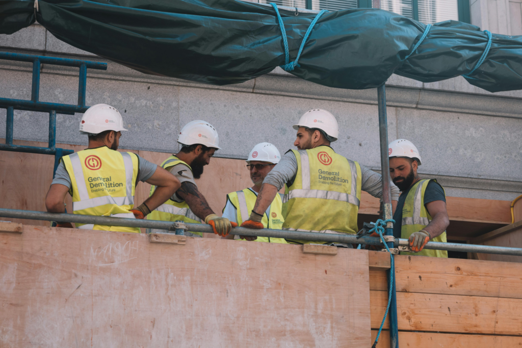Five construction workers in hard hats and high-visibility vests stand together on scaffolding at a worksite.