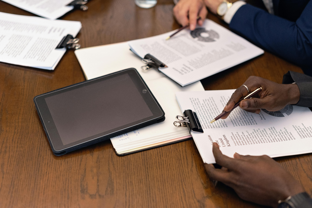 Two professionals review documents on clipboards beside a tablet at a wooden conference table.