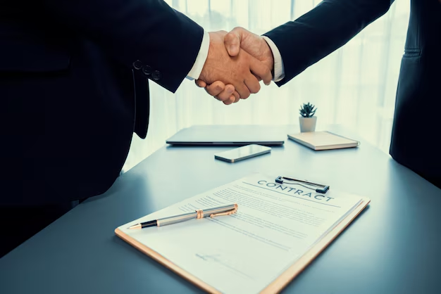Two people in business suits shaking hands over a CONTRACT document and a pen on a desk, representing a written agreement between consenting parties.