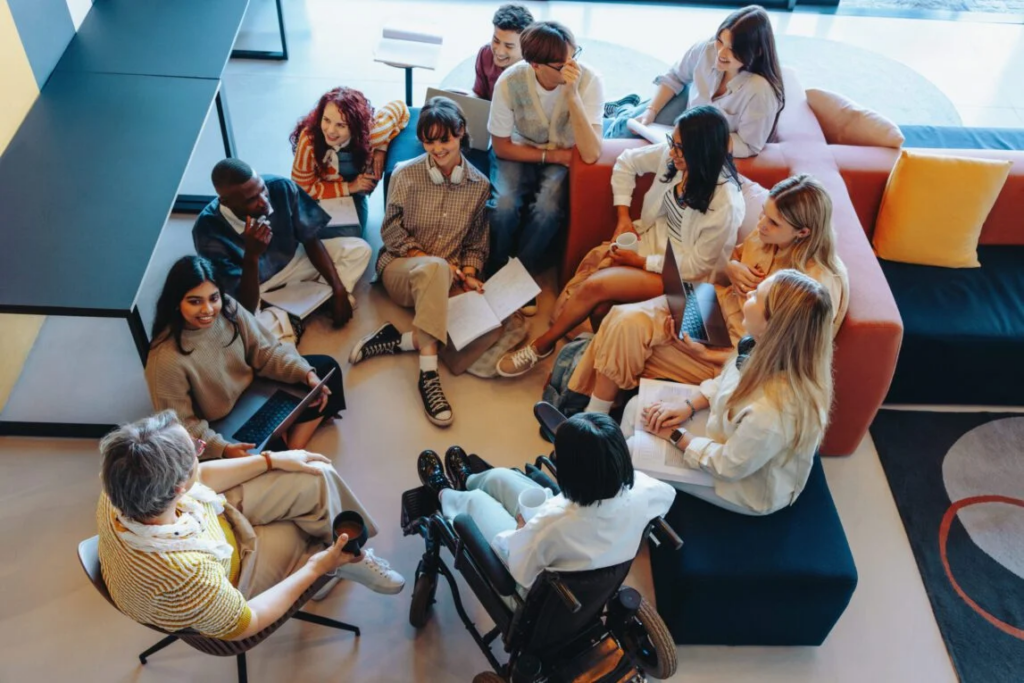 A diverse group of community members, including a person in a wheelchair, gathered in a lounge for an inclusive advocacy discussion.