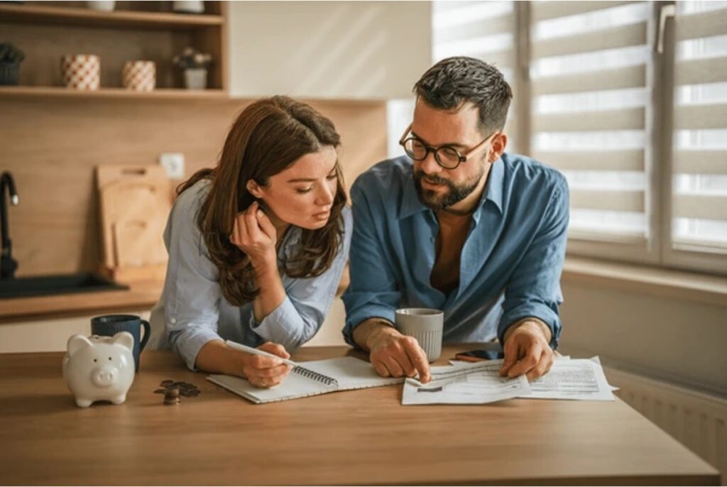 A couple leaning over their kitchen table reviewing estate planning documents, with a piggy bank and loose coins beside them