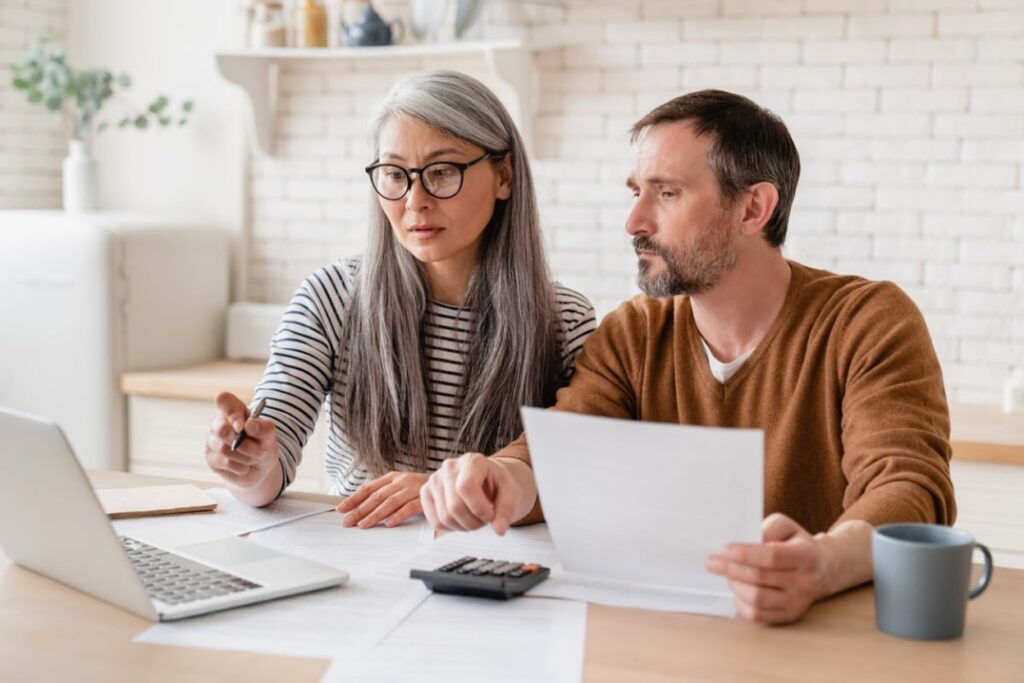 Couple reviewing trust documents with calculator and laptop at kitchen table, discussing estate planning decisions together.