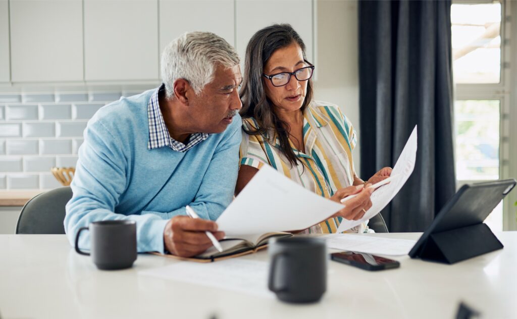 Older couple at kitchen table reviewing documents together, with a tablet and coffee mugs nearby.