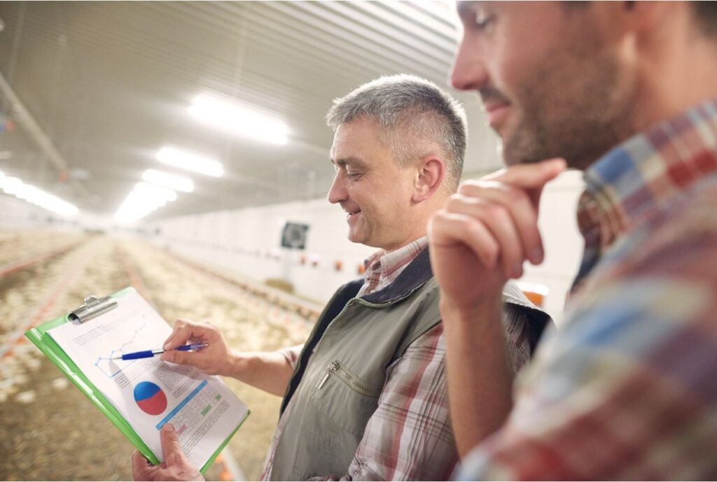 Two farmers reviewing performance charts on a clipboard inside a poultry barn