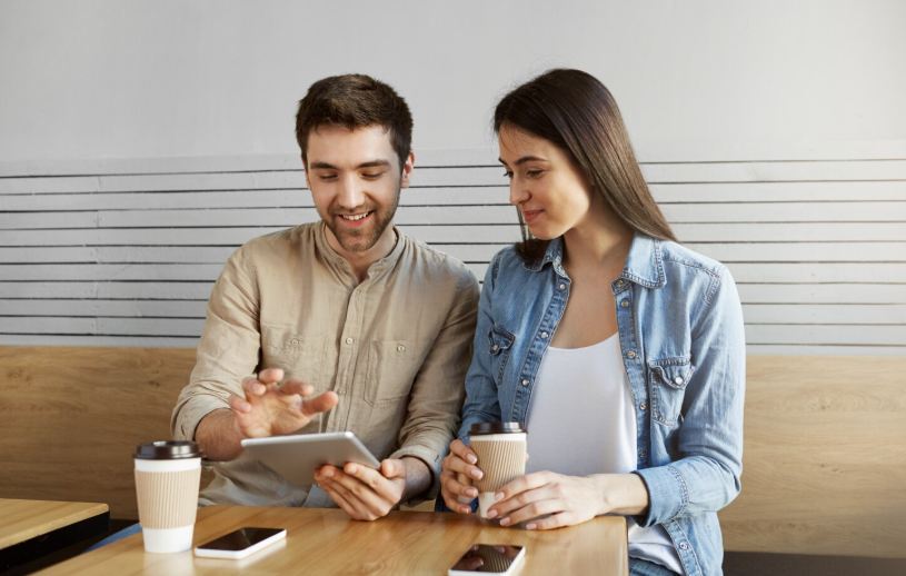 A young couple sitting at a café table looking at a tablet together while drinking coffee.