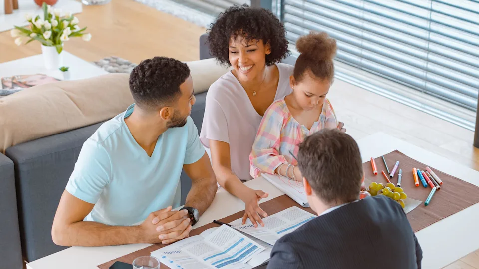 Family with young daughter meeting financial advisor at home, reviewing documents together while discussing long-term financial planning.