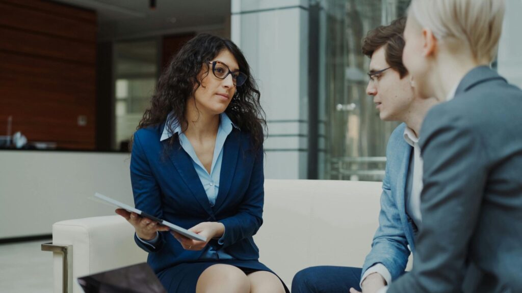 A professional advisor in a navy blazer holds a tablet while speaking with two clients.