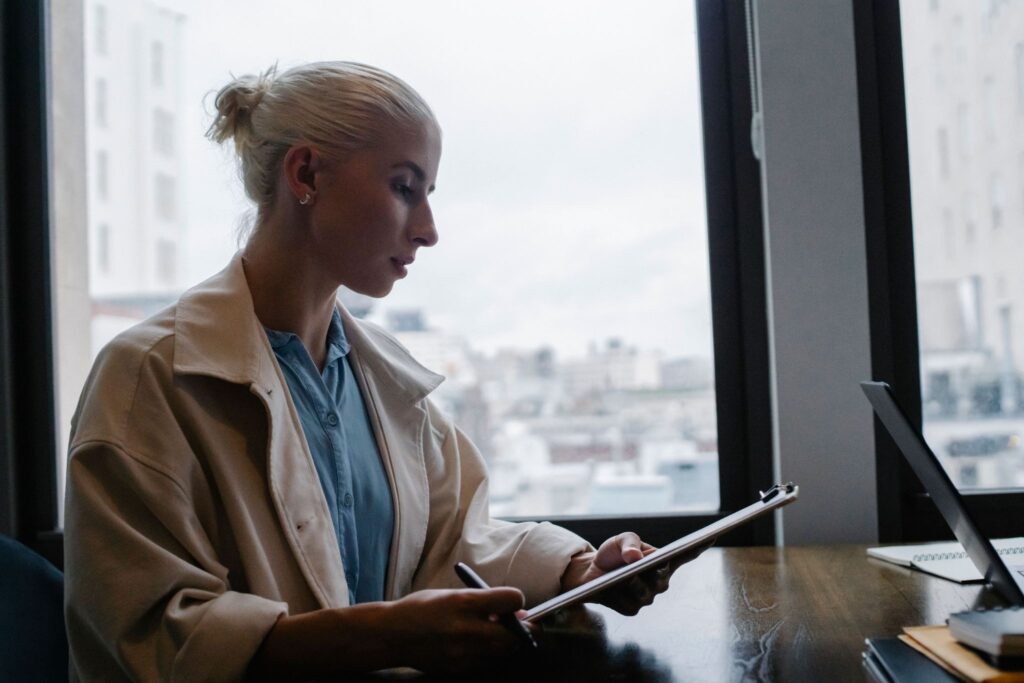 A blonde woman in a tan jacket reviews documents on a clipboard at a desk.